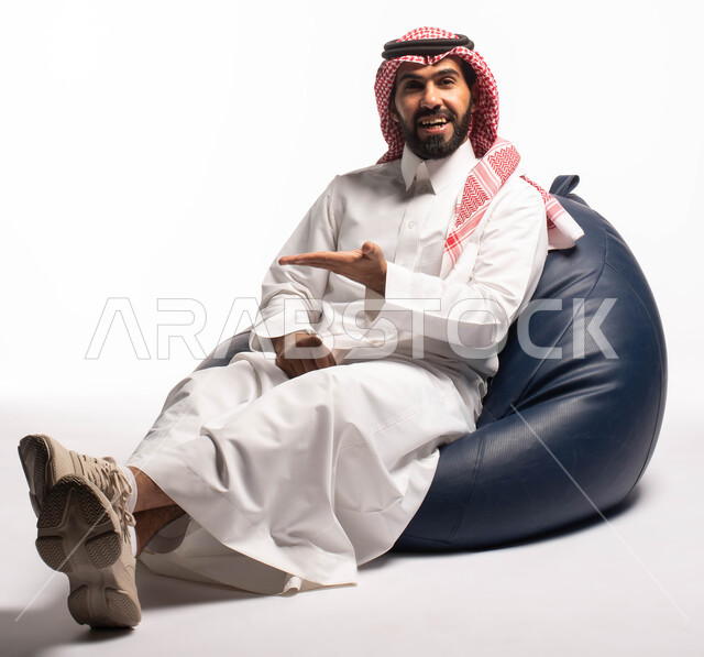 حركات يدين تدل على الإشارة إلى شيء ما, النظر إلى الكاميرا ، بورتريه لرجل عربي سعودي خليجي مبتسم يرتدي الثوب التقليدي يجلس على كرسي مريح, إيماءات وجه تدل على السعادة، خلفيه بيضاء