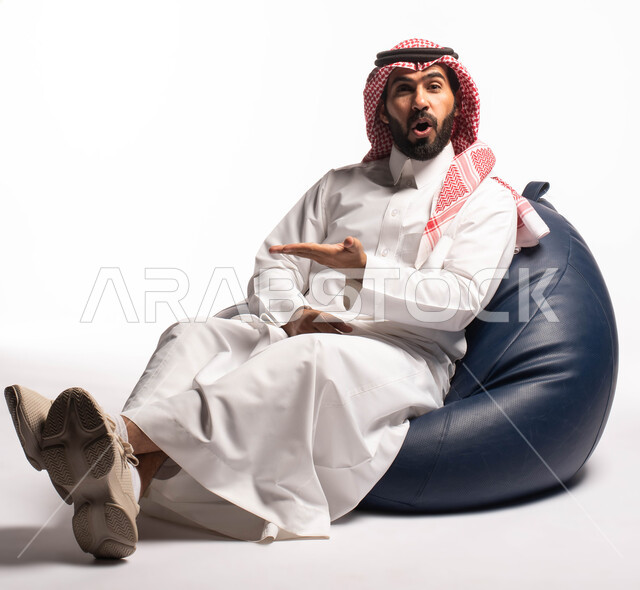 النظر إلى الكاميرا ، بورتريه لرجل عربي سعودي خليجي يرتدي الثوب التقليدي يجلس على كرسي مريح, إيماءات وجه تدل على الدهشة, حركات يدين تدل على الإشارة إلى شيء ما, خلفيه بيضاء