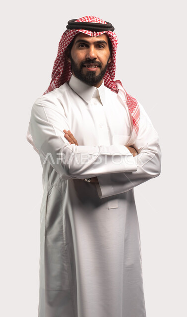 الوقوف بثقة والنظر إلى الكاميرا ، ايماءات وجه تدل على الفرح والسرور ، بورتريه لشاب عربي خليجي سعودي يرتدي الزي السعودي التقليدي يقف متكتف اليدين ، الاهتمام بالمظهر الخارجي ، خلفية بيضاء