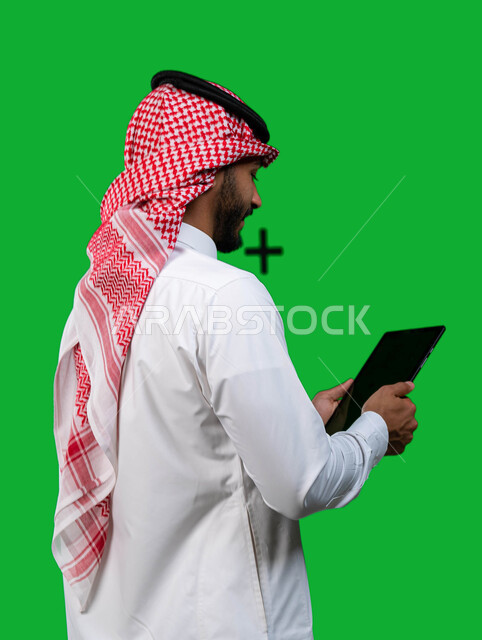 متابعة العمل عن بعد, دمج التقنيه و التكنولوجيا في الحياة العملية, بورتريه من الخلف لرجل أعمال عربي سعودي خليجي يرتدي الشماغ و الثوب التقليدي السعودي يحمل بيده جهاز التابلت, تصفح وسائل التواصل الاجتماعي و الانترنت, كروما، خلفية خضراء. 