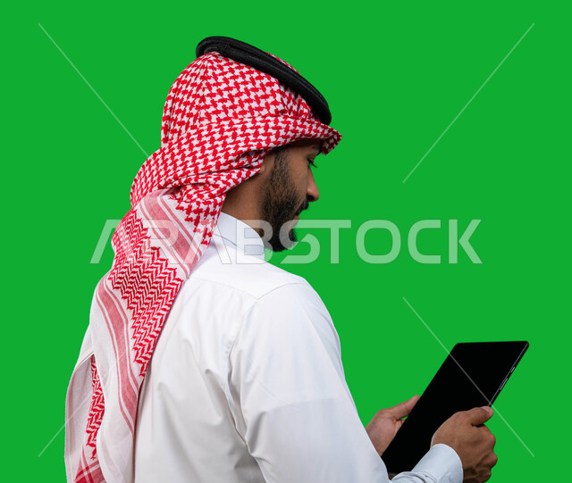Surfing social media and the Internet, following up on remote work, integrating technology and technology into practical life, a portrait from behind of a Saudi Arabian Gulf businessman wearing the traditional Saudi shemagh and thobe holding a tablet in h