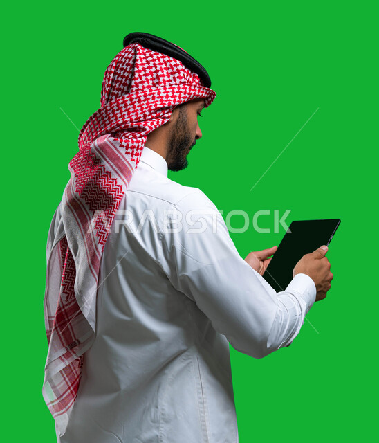 دمج التقنيه و التكنولوجيا في الحياة العملية, متابعة العمل عن بعد, بورتريه من الخلف لرجل أعمال عربي سعودي خليجي يرتدي الشماغ و الثوب التقليدي السعودي يحمل بيده جهاز التابلت, تصفح وسائل التواصل الاجتماعي و الانترنت, كروما، خلفية خضراء. 