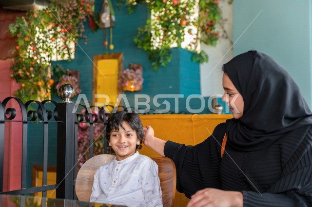 A Saudi Arabian Gulf child spends his time with his mother in the restaurant with gestures and facial expressions that indicate happiness, spending the holiday with the family, a family entertainment atmosphere, unique and distinctive sessions, restaurant