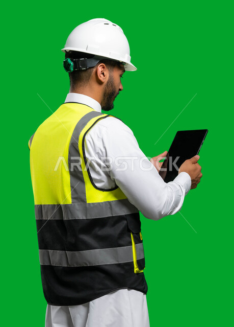 Integration of the practical environment and engineering with technology and technology, electronic engineering plans and designs, a portrait from behind of a Saudi Arabian Gulf engineer wearing a helmet and a protective jacket holding a tablet in his han