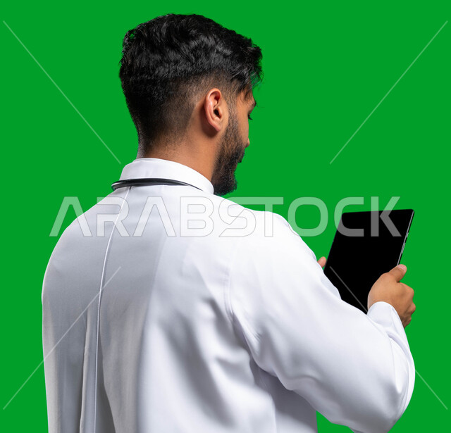 تقديم الاستشارات الطبية عن بعد، بورتريه مقرب من الخلف لطبيب عربي خليجي سعودي يرتدي المعطف الطبي الأبيض, استخدم جهاز التابلت لإعداد التقارير الطبية، دراسة و معاينة الحالة المرضية للمرضى، مفهوم الطب والرعاية الصحية الإلكترونية المنزلية, كروما، خلفية خضراء. 