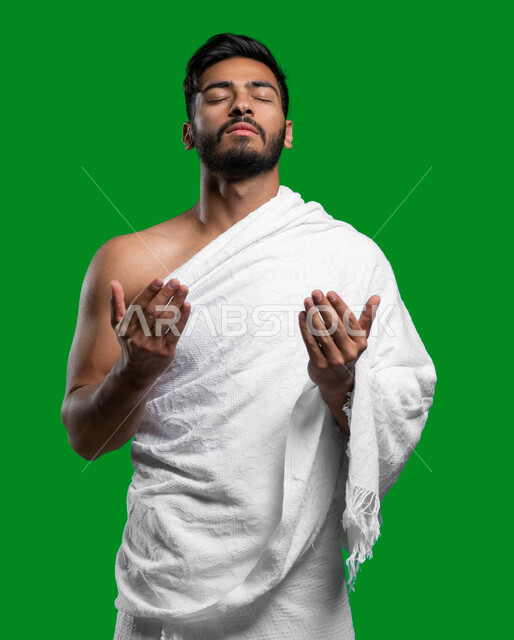 الإجتهاد في العبادة و الحفاظ على الصلاة و التقرب الى الله تعالى,  بورتريه لشاب عربي سعودي خليجي يرتدي ملابس الإحرام يرفع يديه ويدعو الله بإيماءات وجه تدل على التوسل والتضرع لله، فضل العمرة لفي شهر رمضان المبارك, كروما، خلفية خضراء.