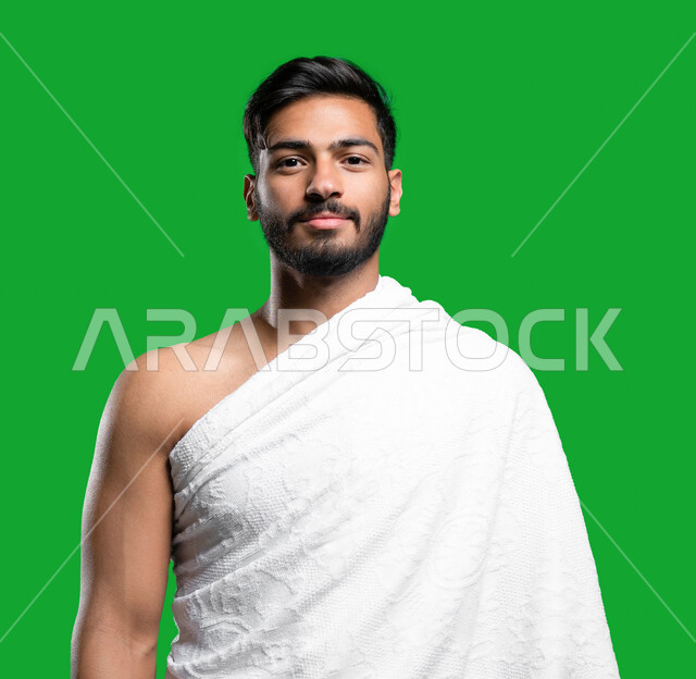 أداء العمرة و التقرب الى الله في شهر رمضان المبارك, بورتريه مقرب لشاب عربي سعودي خليجي مبتسم يرتدي ملابس الإحرام يستعد لأداء مناسك الحج والعمرة ينظر الى الكاميرا، إيماءات وجه تدل على السعادة بزيارة بيت الله الحرام في مكة المكرمة, كروما، خلفية خضراء.