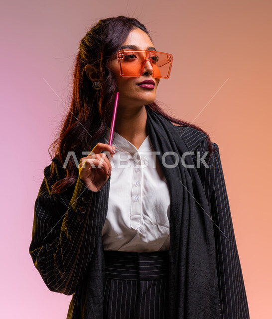 معلمة أنيقة ترتدي نظارات شمسية ملونة عصرية، مهن وأعمال نسائية سعوديه، بورتريه مقرب لسيدة اعمال عربية خليجية سعودية ترتدي اللباس الرسمي تمسك بيدها قلم تقوم بالنظر إلى شيء ما, الاهتمام والاعتناء بالمظهر الخارجي والاناقه، خلفية ملونة