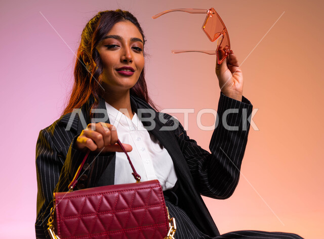 متابعة صيحات الموضة ومواكبة تصاميم الأزياء الحديثة, بورتريه مقرب لشابة عربية خليجية سعودية  مبتسمة ترتدي لباس رسمي عصري حديث وتحمل بيدها حقيبة جلديه ونظارات شمسية ملونة, التسويق للمنتجات السعودية والماركات العالمية الفاخرة,  خلفية ملونة