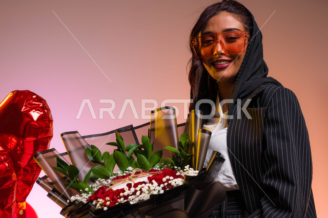 السرور والسعادة أثناء الإحتفال بعيد الحب والمناسبات السعيدة، شراء الزهور الطبيعية والبلالين الحمراء في الفالنتاين، بورتريه مقرب لشابة عربية خليجية سعودية ترتدي النظارات الشمسية و تحمل بيدها باقة ورد جوري أحمر فاخر، خلفية ملونة