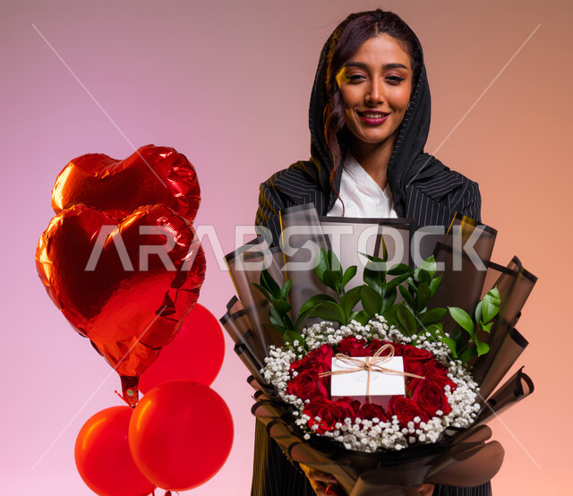 Possession of gifts, balloons and natural flowers on Valentine’s Day, gestures indicating joy and happiness while celebrating holidays and happy occasions, a close-up portrait of a young Saudi Arabian Gulf woman holding in her hand a luxurious red rose bo