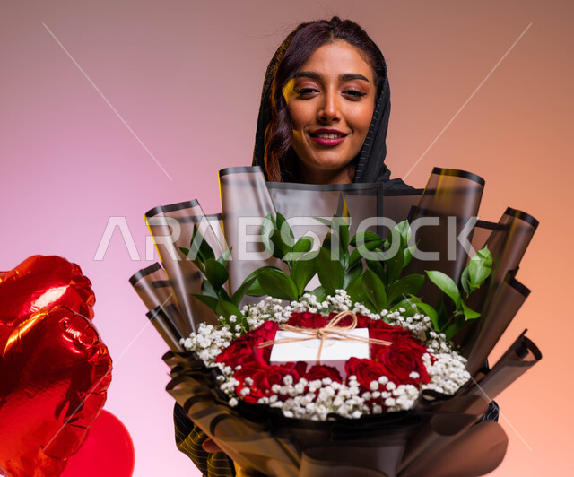 شراء الهدايا و البلالين والزهور الطبيعية في عيد الحب، إيماءات تدل على الفرح والسعادة أثناء الإحتفال بالأعياد و المناسبات السعيدة، بورتريه مقرب لشابة عربية خليجية سعودية تحمل بيدها بوكية ورد جوري أحمر فاخر، خلفية ملونة
