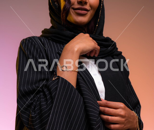 إمرأه مبتسمه ترتدي عباءة سوداء مخططة بأحدث الموديلات والاكسسوارات النسائية السعودية والعالمية، الإهتمام بالموضة والجمال والأناقة، بورتريه مقرب لأيدي شابة عربية سعودية خليجية مزينة بالأساور الذهبيه، خلفية ملونة