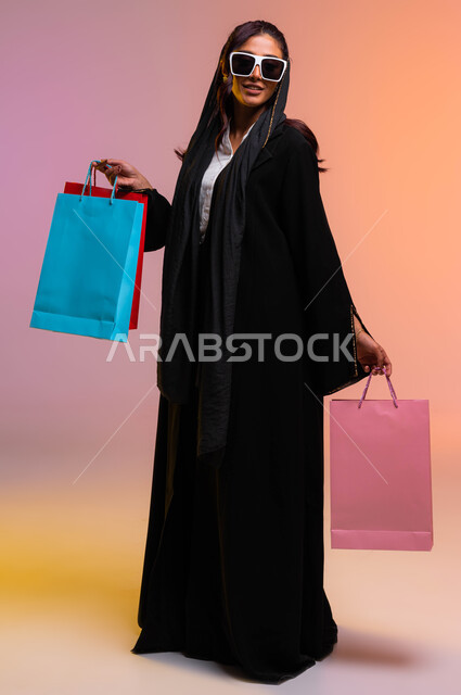 صورة الجسم بالطول الكامل، التسوق الممتع واقتناء المستلزمات والهدايا، بورتريه لامرأة عربية سعودية خليجية ترتدي عباءة سوداء ونظارة شمسية تحمل بيدها أكياس تسوق ملونه، الترويج للمنتجات الفاخرة والاستفادة من العروض والتخفيضات في أسواق السعودية، خلفية ملونة