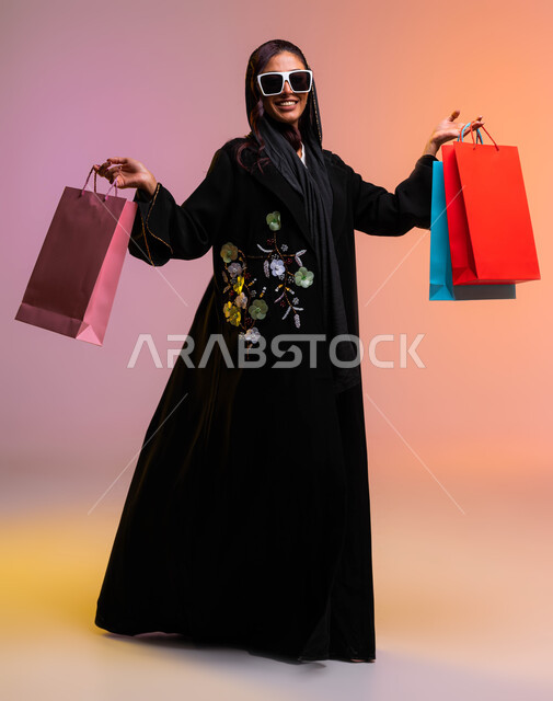 ايماءات السرور بعد التسوق والشراء وتأثيره الإيجابي على المرأة، عروض وتخفيضات موسمية في الأسواق السعودية, بورتريه لامرأة عربية سعودية خليجية مبتسمة ترتدي عباءة سوداء مطرزة ونظارة شمسية تحمل بيدها أكياس تسوق ملونه، صورة الجسم بالطول الكامل، خلفية ملونة
