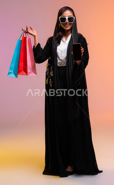 الترويج للمنتجات السعودية، اقتناء المستلزمات والهدايا والتسوق الالكتروني، عروض وتخفيضات موسمية, بورتريه لشابة عربية خليجية سعودية ترتدي عباءة سوداء ونظارات شمسية تحمل بيدها جوال بشاشة سوداء وأكياس التسوق الملونه، صورة الجسم بالطول الكامل، خلفية ملونة