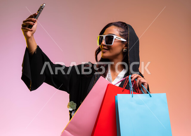 التقاط صور سيلفي تذكارية, مفهوم التصوير والشهرة عبر برامج التواصل الاجتماعي، بورتريه لشابة عربية خليجية سعودية ترتدي لباس عصري حديث ونظارات شمسية تحمل بيدها أكياس التسوق الملونه، التسوق الآمن عبر الانترنت والترويج للمنتجات السعودية الحديثة, خلفية ملونة