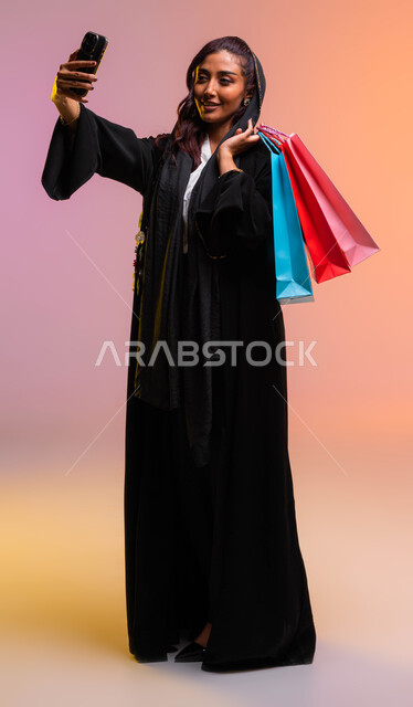 التسوق الآمن عبر الانترنت والترويج للمنتجات السعودية الحديثة, مفهوم التصوير والشهرة عبر برامج التواصل الاجتماعي، التقاط صور سيلفي تذكارية، بورتريه لشابة عربية خليجية سعودية ترتدي لباس عصري حديث تحمل بيدها أكياس التسوق الملونه، خلفية ملونة