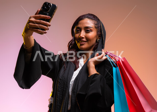 التقاط صورة سيلفي عبر الجوال، مفهوم التصوير والشهرة عبر برامج التواصل الاجتماعي، التسوق من المولات التجارية، بورتريه مقرب لشابة عربية خليجية سعودية ترتدي عباءة سوداء أنيقة تحمل بيدها أكياس التسوق الملونه، خلفية ملونة