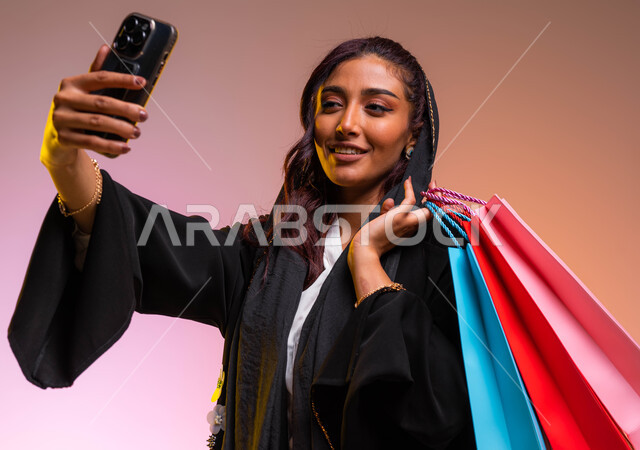 مفهوم التصوير والشهرة عبر برامج التواصل الاجتماعي، التسوق من المولات التجارية،  بورتريه مقرب لشابة عربية خليجية سعودية ترتدي عباءة سوداء أنيقة تحمل بيدها أكياس التسوق الملونه، التقاط صورة سيلفي عبر الجوال، خلفية ملونة