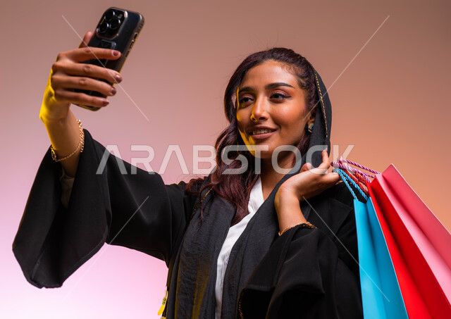 التسوق من المولات التجارية، مفهوم التصوير والشهرة عبر برامج التواصل الاجتماعي، بورتريه مقرب لشابة عربية خليجية سعودية ترتدي عباءة سوداء أنيقة تحمل بيدها أكياس التسوق الملونه، التقاط صورة سيلفي عبر الجوال، خلفية ملونة