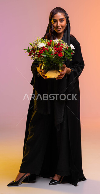 صورة الجسم بالطول الكامل, تنسيق باقات الزهور في الأعياد والمناسبات السعيدة, متاجر ومحلات بيع وشراء الأزهار الطبيعية, بورتريه لأنثى عربية خليجية سعودية مبتسمة ترتدي عباءة سوداء تحمل بيدها مزهرية ورد أحمر فاخر، خلفية ملونة