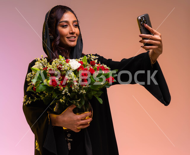 ورود وزهور طبيعية جميلة للأعياد والمناسبات, ايماءات تدل على الفرح والسعاده, محلات ومتاجر بيع وشراء الأزهار, بورتريه مقرب لامرأة عربية خليجية سعودية مبتسمة ترتدي عباءة سوداء تحمل بيدها حوض ورد أحمر فاخر تلتقط صور سيلفي تذكارية عبر الجوال, خلفية ملونة