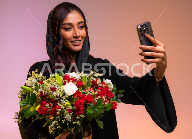 ايماءات تدل على الفرح والسعاده, محلات ومتاجر بيع وشراء الأزهار, ورود وزهور طبيعية جميلة للأعياد والمناسبات, بورتريه مقرب لامرأة عربية خليجية سعودية مبتسمة ترتدي عباءة سوداء تحمل بيدها حوض ورد أحمر فاخر تلتقط صور سيلفي تذكارية عبر الجوال, خلفية ملونة