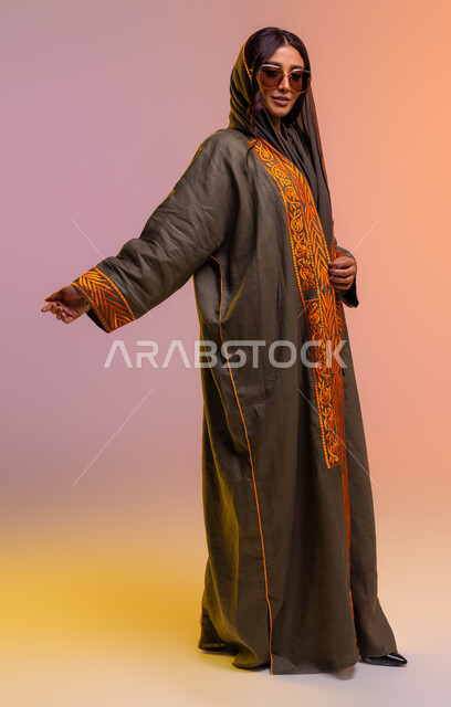 صورة الجسم بالطول الكامل، ايماءات الأنوثة والجمال, عرض الأزياء بالتصاميم والموديلات الحديثة، بورتريه لامرأة عربية خليجية سعودية ترتدي عباءة بنية مطرزة ونظارات شمسية عصرية تقف بثقة، مفهوم الموضة والأناقة، خلفية ملونة