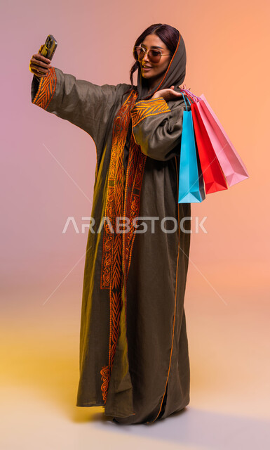 التبضع و التسوق من المولات التجارية، مفهوم التصوير والشهرة عبر وسائل التواصل الاجتماعي، التقاط صور السيلفي باستخدام الهاتف، بورتريه لسيدة عربية خليجية سعودية ترتدي عباءة مطرزة ونظارة شمسية تحمل أكياس تسوق ملونه، صورة الجسم بالطول الكامل، خلفية ملونة