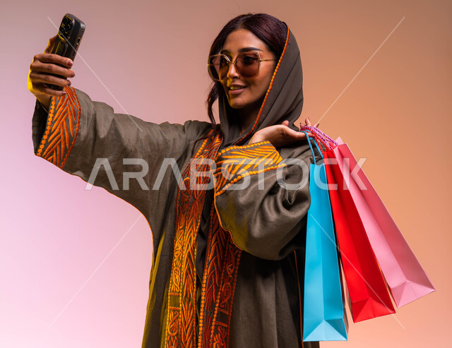  التسوق الالكتروني الآمن عبر الانترنت، بورتريه مقرب  لشابة عربية خليجية سعودية مبتسمة ترتدي عباءة بنيه مطرزة ونظارات شمسية عصرية تحمل بيدها أكياس التسوق الملونه تقوم بالتقاط صور سيلفي، مشاركة اليوميات عبر وسائل التواصل الاجتماعي، خلفية ملونة