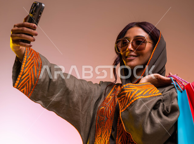 مشاركة اليوميات عبر وسائل التواصل الاجتماعي، بورتريه مقرب  لشابة عربية خليجية سعودية مبتسمة ترتدي عباءة بنيه مطرزة ونظارات شمسية عصرية تحمل بيدها أكياس التسوق الملونه تقوم بالتقاط صور سيلفي، التسوق الالكتروني الآمن عبر الانترنت، خلفية ملونة