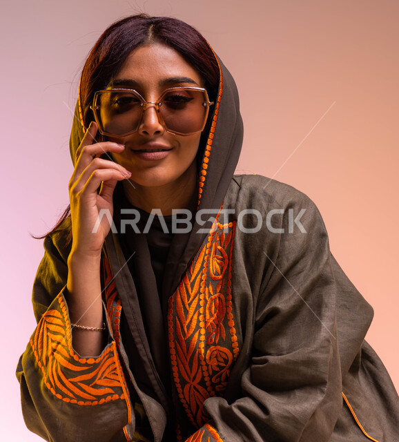  الاهتمام بمتابعة صيحات الموضة, ملابس واكسسوارات نسائية بتصاميم معاصرة, مفهوم الجمال والأناقة, بورتريه مقرب لامرأه عربية خليجية سعودية مبتسمة ترتدي عباءة بنيه مطرزة ونظارات شمسية عصرية, ايماءات تدل على التفكير والتأمل, خلفية ملونة