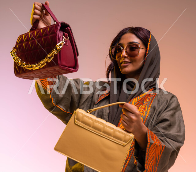  ايماءات الإعجاب والسعادة لامتلاك حقيبه جديدة، قضاء وقت ممتع في اختيار الحقائب، تسوق وشراء المنتجات السعوديه والماركات العالمية الفاخرة، بورتريه مقرب لإمرأة عربية خليجية سعودية ترتدي عباءة عصرية مطرزة ونظارات شمسية تحمل بيدها حقائب أنيقة، خلفية ملونة