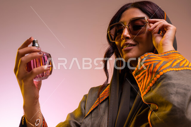 استخدام عطور وزيوت عطرية طبيعية، التسويق لمنتجات العطور الخليجيه والعالمية، بورتريه مقرب لإمرأه عربيه سعودية خليجية مبتسمة ترتدي عباءة مطرزة ونظارات شمسية تقوم برش العطر، مفهوم الأناقة والإهتمام بالمظهر الخارجي، التطيب والتعطر بالعود العطري، خلفية ملونة