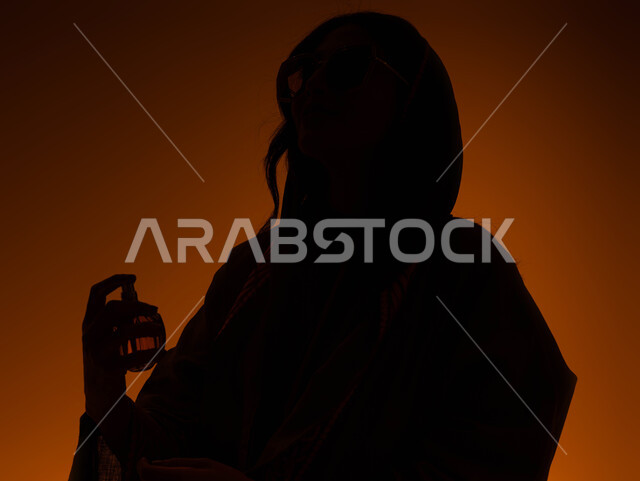 مفهوم الأناقة والإهتمام بالمظهر الخارجي، التسويق لمنتجات العطور النسائيه الخليجيه، بورتريه صورة ظلية مقربة لإمرأه عربيه سعودية خليجية تقوم برش العطر، استخدام الزيوت العطرية الطبيعية، التطيب والتعطر بالعود العطري، خلفية ملونة، سيلوليت