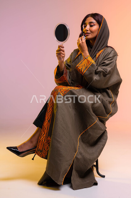 دورات تعليمية لمكياج تتوريال، الإهتمام بالجمال والبشرة وإبراز الانوثة، متجر بيع مستحضرات التجميل، بورتريه لإمرأة عربية خليجية سعودية ترتدي عباءة بنية مطرزة تحمل مرآة وتقوم بوضع أحمر شفاه، تسويق لمنتجات المكياج السعودية و الماركات العالمية, خلفيه ملونه