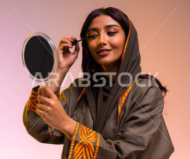 مكياج تتوريال وتسويق لمنتجات سعودية وماركات عالمية فاخرة، خبيرة مستحضرات التجميل النسائية، بورتريه مقرب لإمرأة عربية خليجية سعودية ترتدي عباءة بنية مطرزة تحمل مرآة تقوم بوضع المسكارا لإبراز العين، مفهوم الإهتمام والعناية بالجمال والبشرة, خلفيه ملونه