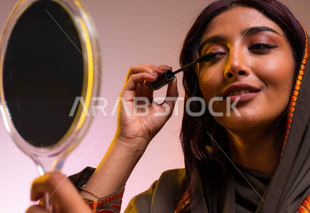 خبيرة مستحضرات التجميل النسائية، بورتريه مقرب لإمرأة عربية خليجية سعودية ترتدي عباءة بنية مطرزة تحمل مرآة تقوم بوضع المسكارا لإبراز العين، دورات تعليمية لمكياج تتوريال, تسويق لمنتجات سعودية وماركات عالمية فاخرة، مفهوم الإهتمام بالجمال والبشرة, خلفيه ملونه