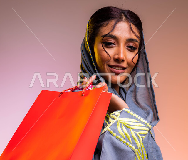 عروضات وتخفيضات موسمية في الأسواق السعودية، ايماءات السرور بعد التسوق والشراء وتأثيره الإيجابي على المرأة, بورتريه مقر لامرأة عربية سعودية خليجية مبتسمة ترتدي عباءة رمادية مطرزة تنظر الى الكاميرا تحمل بيدها أكياس تسوق ملونه، خلفية ملونة