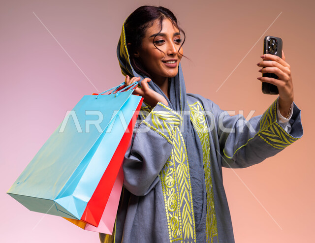 التسوق من المحلات والمراكز التجارية الالكترونية، التقاط صور سيلفي باستخدام الهاتف المحمول، مفهوم التصوير والشهرة عبر وسائل التواصل الاجتماعي، بورتريه مقرب لشابة سعودية خليجية ترتدي عباءة رمادية مطرزة تحمل أكياس تسوق ملونة، خلفية ملونة.