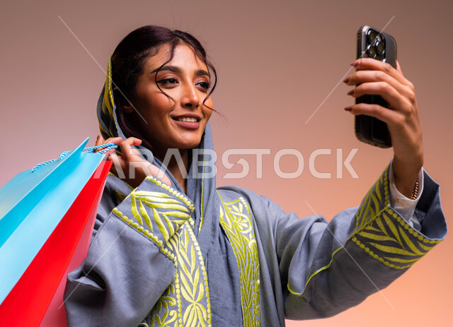 التسوق الالكتروني من المحلات والمراكز التجارية، التقاط صور سيلفي باستخدام الهاتف المحمول، مفهوم التصوير والشهرة عبر وسائل التواصل الاجتماعي، بورتريه مقرب لشابة سعودية خليجية ترتدي عباءة رمادية مطرزة تحمل أكياس تسوق ملونة، خلفية ملونة.