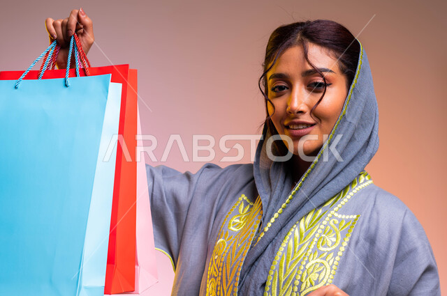 اقتناء بعض المستلزمات والهدايا، ايماءات الإعجاب والاستمتاع بالتسوق وشراء الحقائب الجديدة، الترويج للمنتجات السعوديه و الماركات العالمية الفاخرة، بورتريه مقرب لإمرأة عربية سعودية خليجية ترتدي عباءة رمادية مطرزة تحمل بيدها أكياس تسوق ملونه، خلفية ملونة
