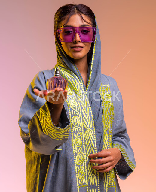 التسويق لمنتجات سعودية وعالمية, التجمل والتزيين النسائي, بورتريه لامرأة عربية سعودية خليجية ترتدي عباءة مطرزة ونظارة شمسية تحمل بيدها زجاجة عطر عصرية, إيماءات السعادة لشراء زيوت عطرية جديدة, خلفية ملونة