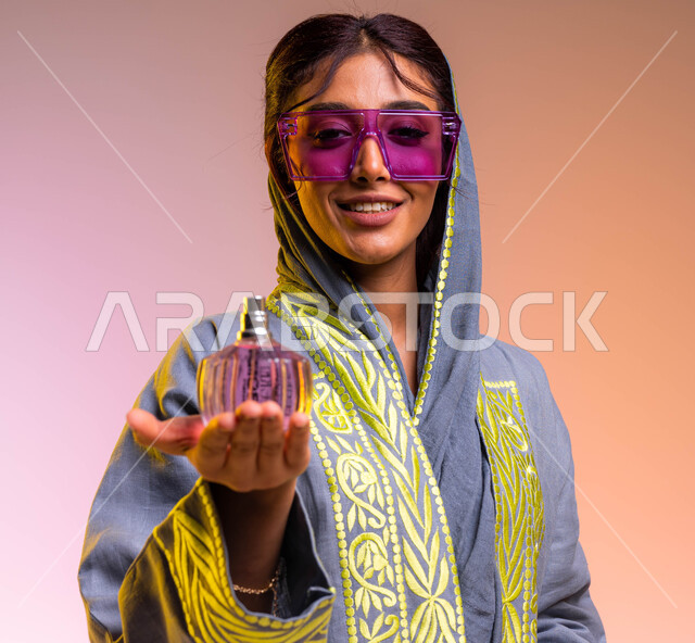 التجمل والتزيين النسائي, بورتريه مقرب لامرأة عربية سعودية خليجية ترتدي عباءة مطرزة ونظارة شمسية تحمل بيدها زجاجة عطر عصرية, إيماءات السعادة لشراء زيوت عطرية جديدة, التسويق لمنتجات سعودية وعالمية، خلفية ملونة