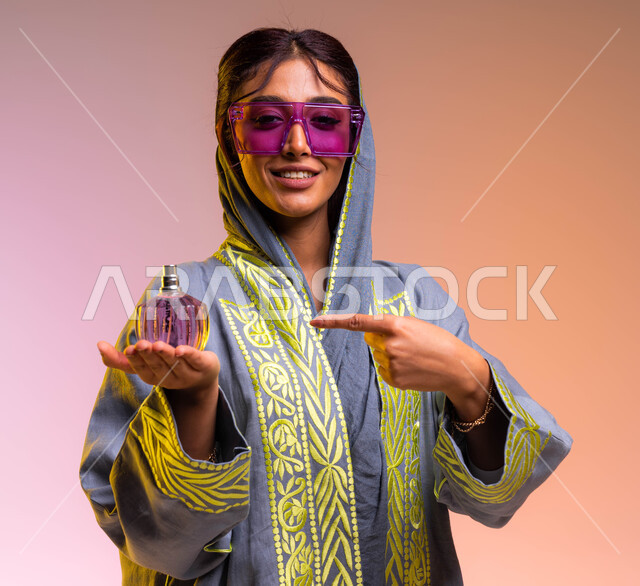 إيماءات السعادة لشراء زيوت عطرية جديدة, التجمل والتزيين النسائي, بورتريه لامرأة عربية سعودية خليجية ترتدي عباءة مطرزة ونظارة شمسية تشير إلى زجاجة عطر عصرية, التسويق لمنتجات العطور السعودية والعالمية, خلفية ملونة