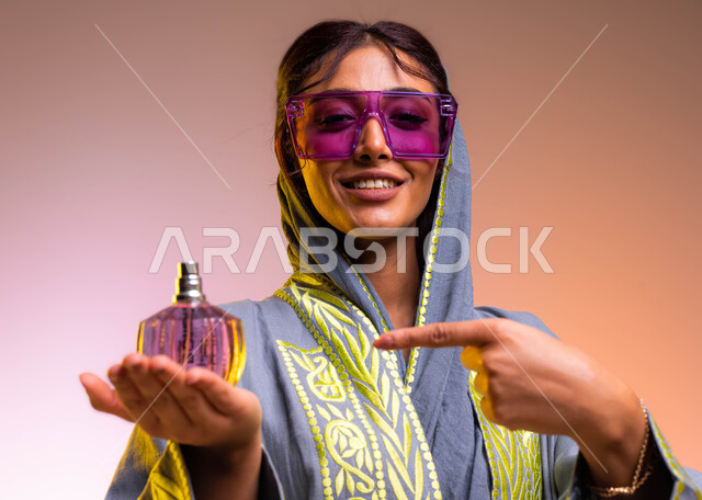 التسويق لمنتجات العطور السعودية والعالمية, التجمل والتزيين النسائي, بورتريه لامرأة عربية سعودية خليجية ترتدي عباءة مطرزة ونظارة شمسية تشير إلى زجاجة عطر عصرية, إيماءات السعادة لشراء زيوت عطرية جديدة, خلفية ملونة