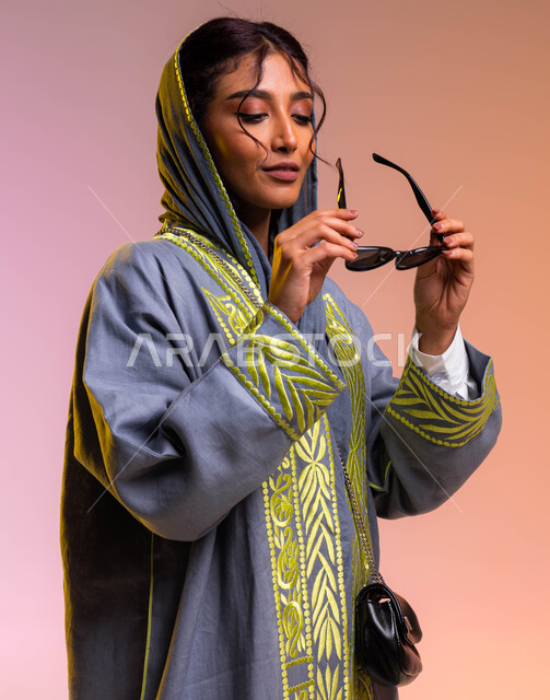 الاستمتاع بإمتلاك اكسسوارات عصرية, مفهوم الموضة والأناقة, بورتريه لإمرأة عربية خليجية سعودية تقوم بتجربة النظارات الشمسية الجديدة, إيماءات تدل على الانوثة والجمال, إرتداء عباءة رمادية مطرزة أنيقة وحقيبة جلد سوداء معاصرة, خلفية ملونة