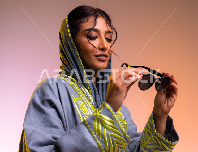 إيماءات تدل على الانوثة والجمال, مفهوم الموضة والأناقة, بورتريه مقرب لإمرأة عربية خليجية سعودية تقوم بتجربة النظارات الشمسية الجديدة, الاستمتاع بإمتلاك اكسسوارات عصرية, إرتداء عباءة رمادية مطرزة أنيقة, خلفية ملونة
