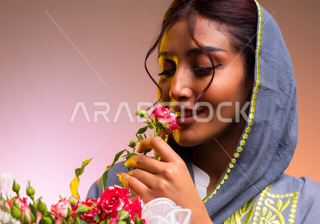 إيماءات الإعجاب بالروائح العطرية الزكية للورد، محلات ومتاجر بيع وشراء الزهور الطبيعية، امتلاك باقات الورود والهدايا في الأعياد والمناسبات السعيدة، بورتريه مقرب لشابة عربية خليجية سعودية ترتدي عباءة مطرزة عصرية تقوم بشم رائحة الزهور، خلفية ملونة